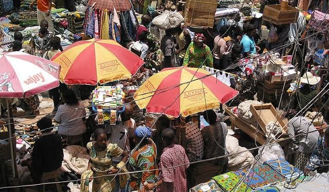 aerial view of a lot of people in close proximity in a market area. There are two big umbrellas close to each other with people under it, shading themselves from the heat of the sun