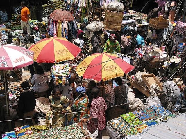 aerial view of a lot of people in close proximity in a market area. There are two big umbrellas close to each other with people under it, shading themselves from the heat of the sun