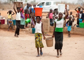 2 girls carrying water on their head and holding one bucket of water in their hands while supporting the water on their head with the other hand. People in the background are also fetching water
