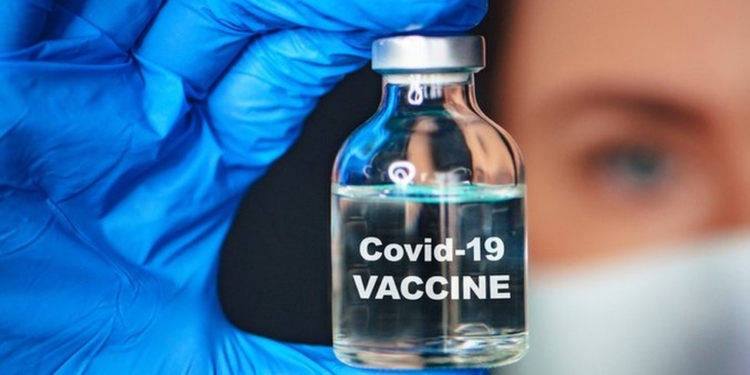 A gloved hand holding a transparent bottle (with the middle finger at the top and thumb at the bottom) containing a liquid in it. Covid-19 vaccine is written on the bottle