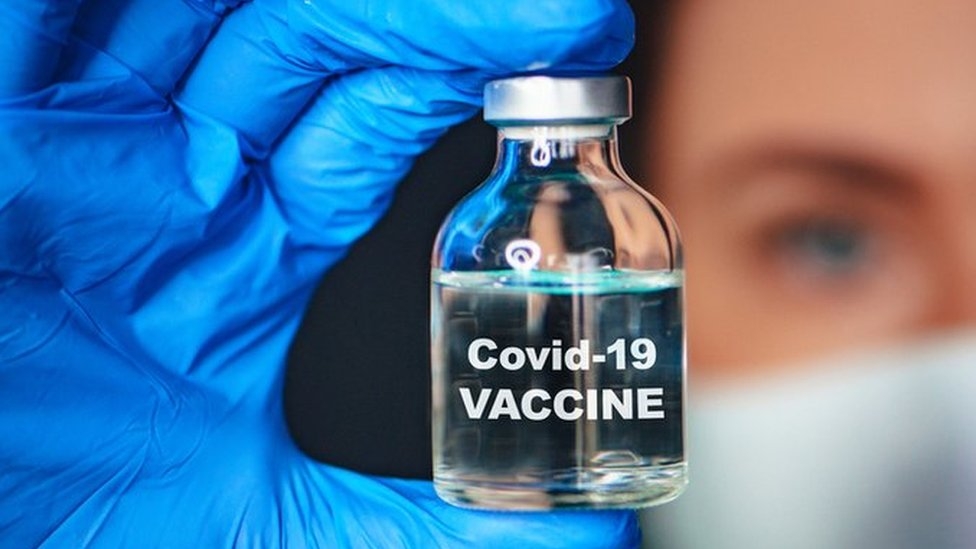 A gloved hand holding a transparent bottle (with the middle finger at the top and thumb at the bottom) containing a liquid in it. Covid-19 vaccine is written on the bottle