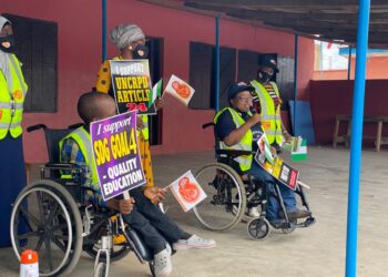 Two people sitting on a wheel chair and two people standing, all of them are holding flags and placards to advocate for quality and inclusive education