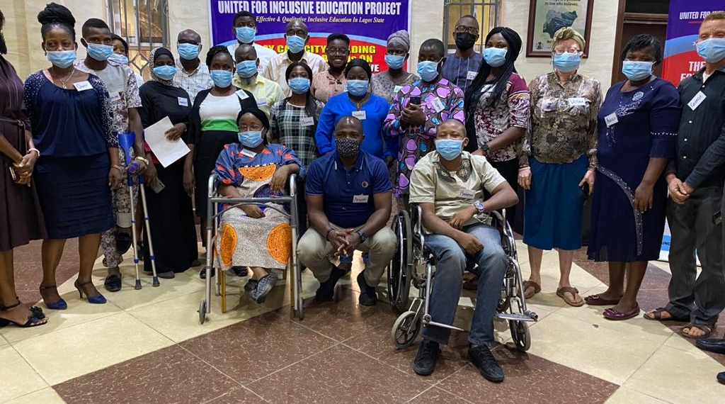 People standing together in a group picture, there people in front. There is one wheelchair user, a walking aide and someone squatting