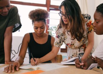 Women standing together and writing on a table, one of them is seated, there are three women in total