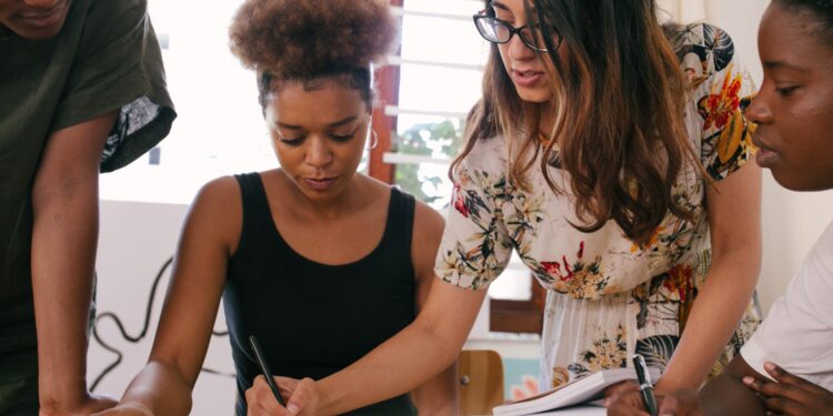 Women standing together and writing on a table, one of them is seated, there are three women in total
