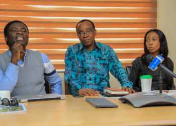 2 men and one woman seated, microphone in front of them, on a table