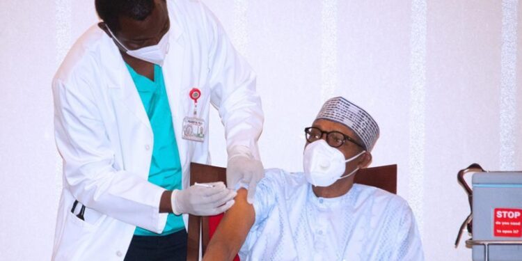 President Buhari sitting down while the medical officer is standing and holding an injection to vaccinate the president