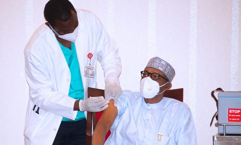President Buhari sitting down while the medical officer is standing and holding an injection to vaccinate the president