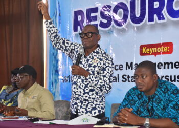 A picture of four men indoors, three seated and one standing talking with a microphone and his right hand up.