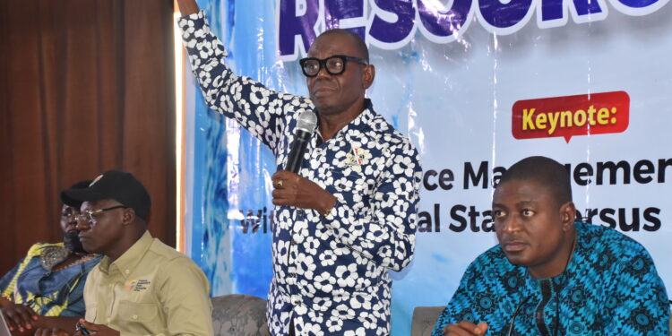 A picture of four men indoors, three seated and one standing talking with a microphone and his right hand up.
