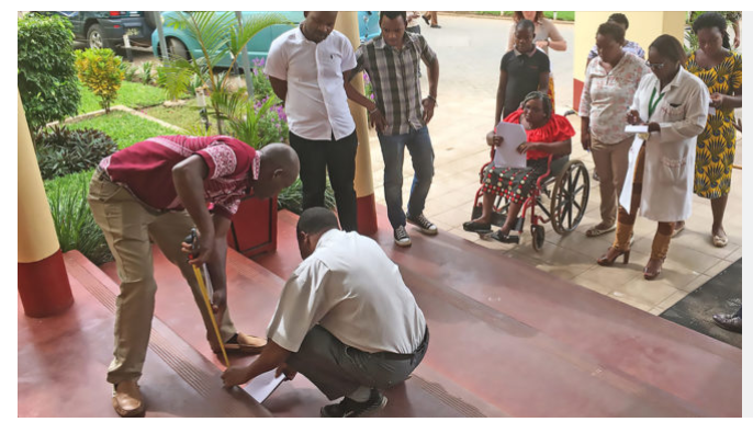 A woman on wheelchair moving towards a building with staircase and there are people standing by the staircase and beside her