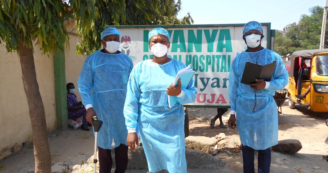In the picture, there are three persons standing out door kitted in COVID19 protective gears