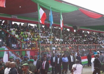 An image of a lot of people gathered outdoor. Some standing others sitting under a shade.