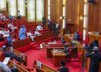 Nigerian senators during a plenary session indoor. Some persons are sitting and others are standing. The senate president is seated on the right hand side on an elevated platform facing the members of senate