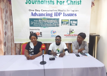 Three persons, a woman on the left, a man in the centre and another woman on the right sitting at a high table indoors. Their hands are on the table and hehind them is a banner of Journalist for Christ with donor logos