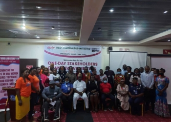 Group of persons standing next to eah other and others sitting next to each other indoor. Behind is a banner with the inscription deaf women aloud initiative