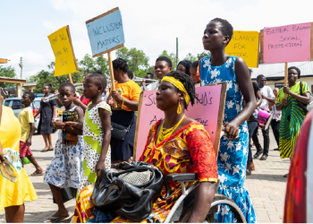 A woman on wheel chair in a rallying crowd outdoor. She is supported from behind by another woman. The people in the crowd are carrying placards - one reads "inclusion matters"