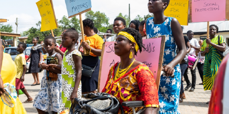 A woman on wheel chair in a rallying crowd outdoor. She is supported from behind by another woman. The people in the crowd are carrying placards - one reads "inclusion matters"