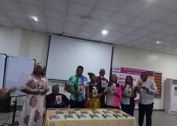 Eight persons standing in front of a table indoors holding books in their hands. Books are also lined up on the table