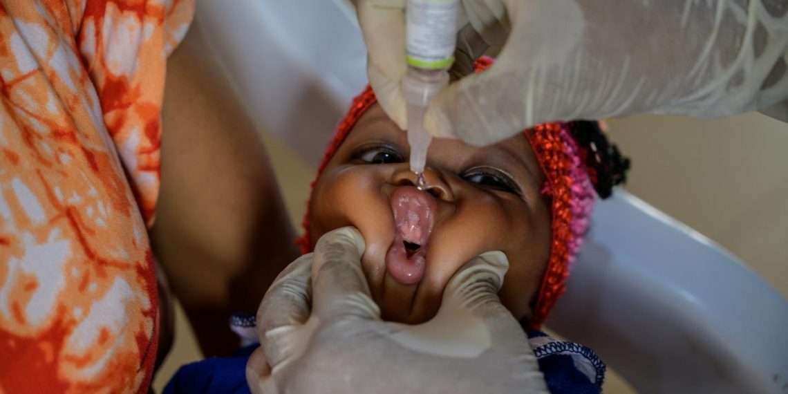 A baby's mouth is gently pressed with a gloved left hand and the right gloved hand is holding a vaccine pointing too the mouth of the baby. The baby's head is resting on the lap of a woman