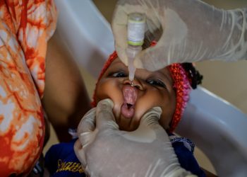 A baby's mouth is gently pressed with a gloved left hand and the right gloved hand is holding a vaccine pointing too the mouth of the baby. The baby's head is resting on the lap of a woman