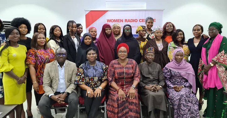 People in a group picture, some are seated while some are standing. There is a banner at the background giving a description of the Women Radio Centre