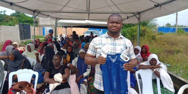 A man standing outdoor under a canopy holding a blue sack in his hand. Some women are seated under the canopy behind him