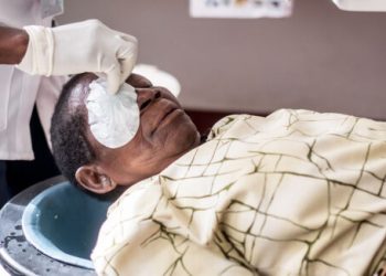 A woman lying down has her eye bandages removed by a hand with gloves