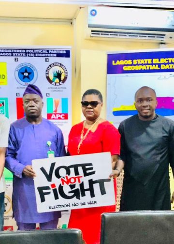 Eight people standing indoor. Two of them have a placard in their hands that reads "vote not fight"