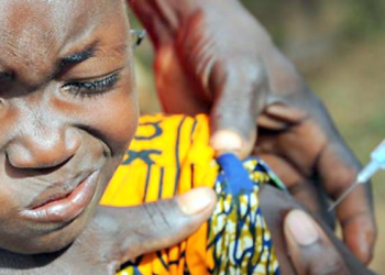 Image of child receiving injection on the left arm. Two hands acting as support to hold the child's shirt from falling