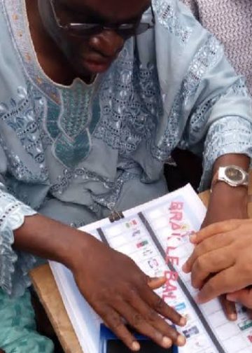 A man sitting outdoor. He has both hands on a ballot paper. A hand of someonr else is on the man's left hand as support