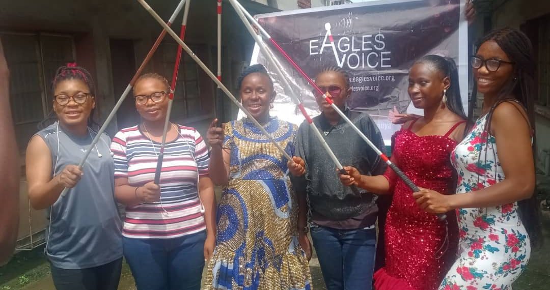5 women holding their white cane in a group picture. There is a banner in the backgroud. he white canes were raised.