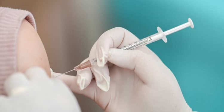 A hand with surgical gloves holding an injection pointing at an arm