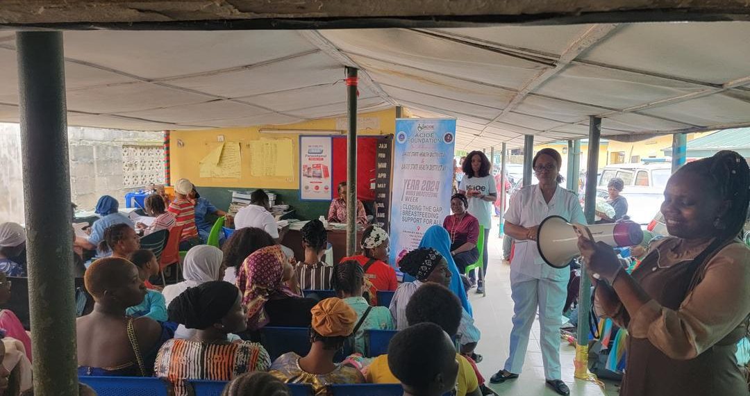 A group of people sitting indoor. One lady is standing holding a megaphone. Two other ladies are also standing in the room.