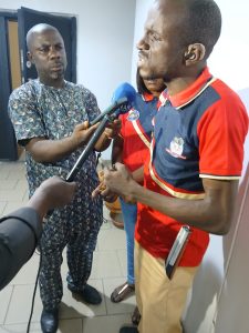 A man standing indoor in front of two people with a microphone in their hand