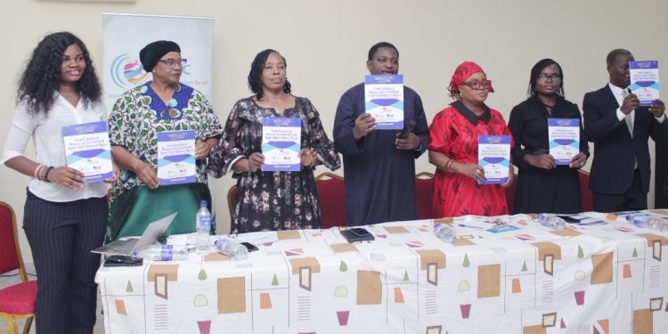 Seven people standing behind a long table indoor with each of them holding a book up to their chest level