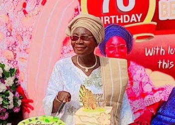 A woman staning out door. There are two cakes on a table in front of her