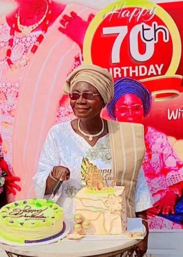 A woman staning out door. There are two cakes on a table in front of her