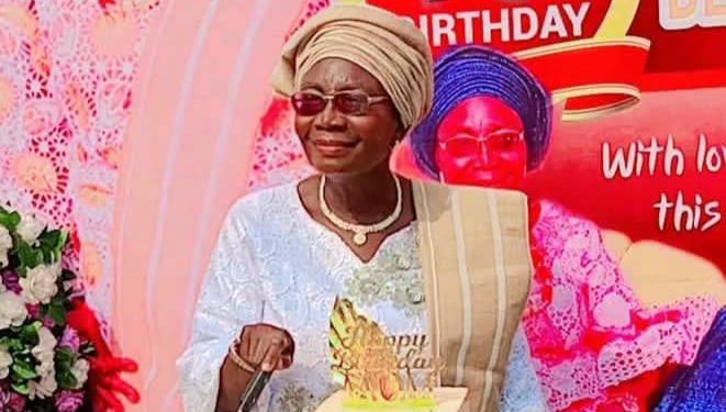 A woman staning out door. There are two cakes on a table in front of her