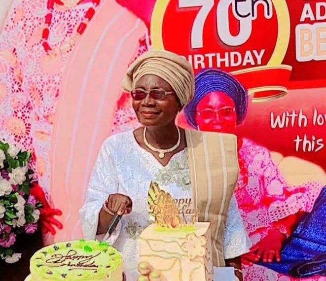 A woman staning out door. There are two cakes on a table in front of her