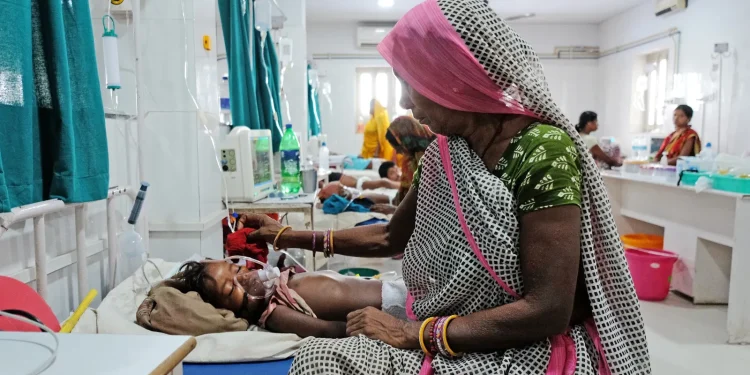 A woman sitting on one side of a bed in the hospital. Her right hand is raised above an infant who is sleeping on the bed covered with an oxygen mask (indoor)