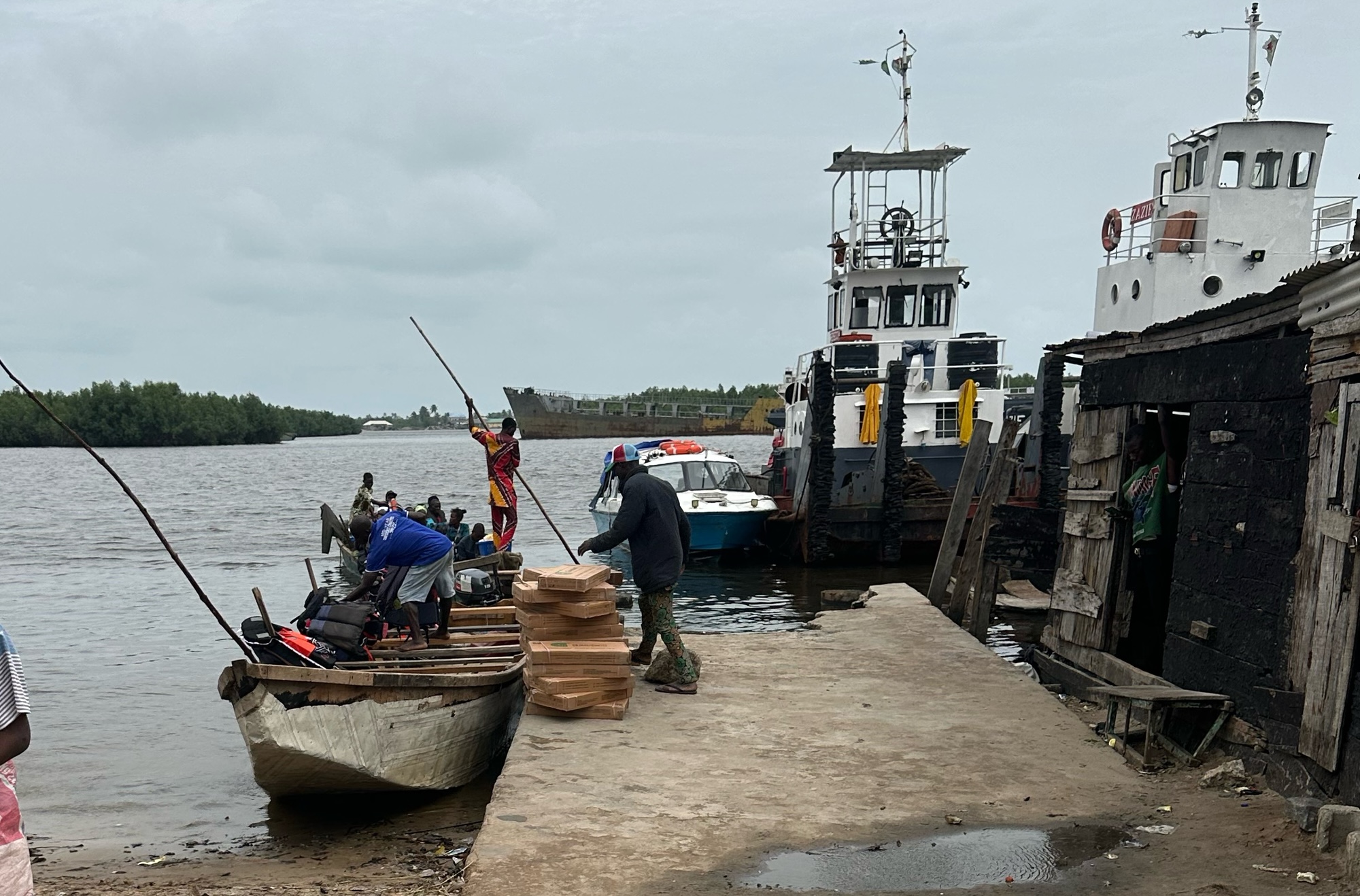 A concrete walkway leading to a large body of water. To the left side is a boat and to the left is a building made of wood. Up ahead is a bigger boat on the water