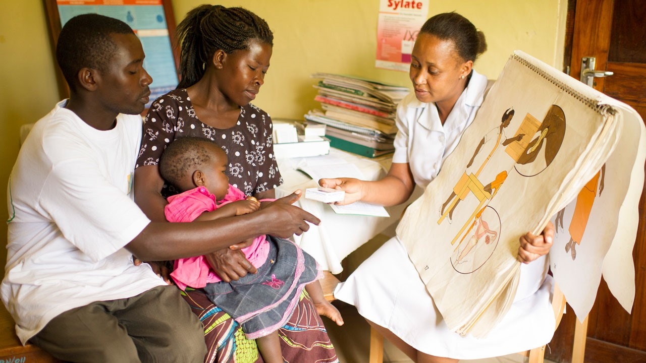 Image of a man and two women indoor. One of the women is holding a child in her hand. The other woman is holding a poster in her hand. All of them are indoor