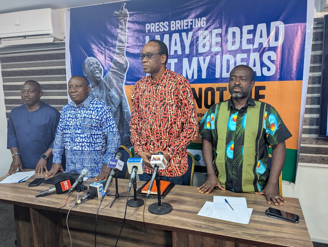 Four men standing indoors. In front of them is a table with six microphones on it
