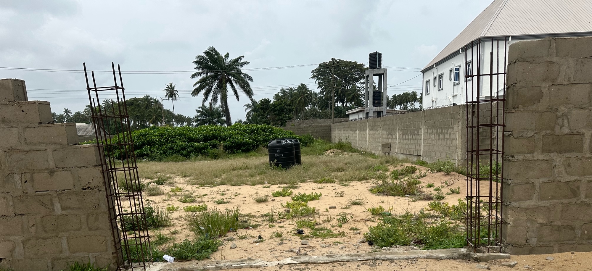 A fenced land with a big plastic tank at one side of the fence. The entrance gate to the land is not completely built. It has metal rods showing on both sides of the entrance