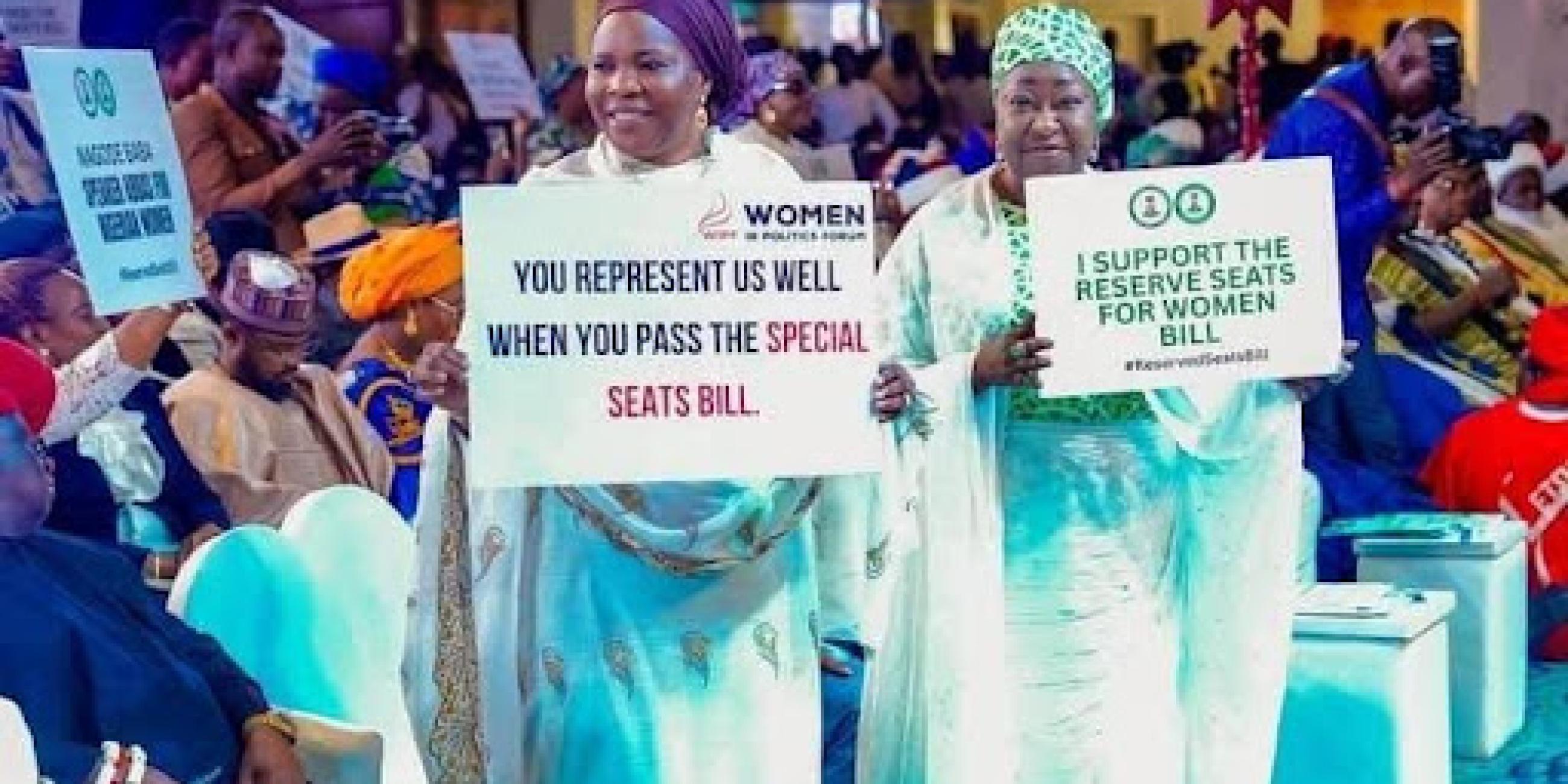 Two women standing indoor holding a placard in a room full of people