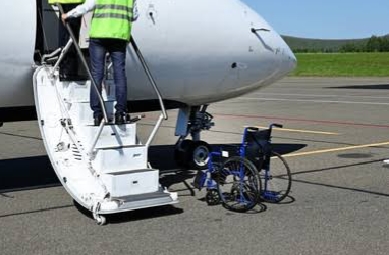 Front view of a parked plane with door open. A person is standing on the stairs that leads to the plane through the door. At the foot of the stairs is an empty wheelchair
