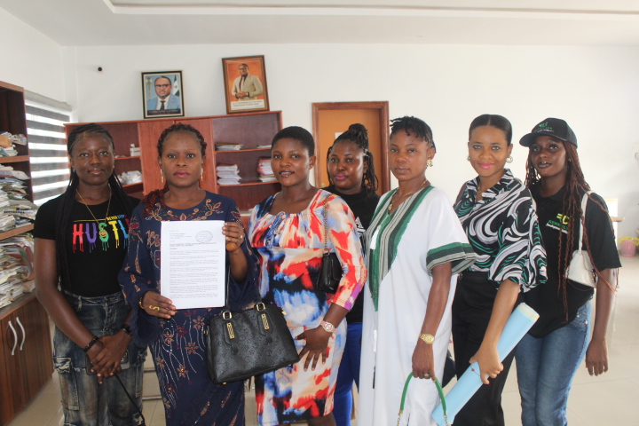 Seven women standing indoors. One of them is holding a paper document with both hands to her chest
