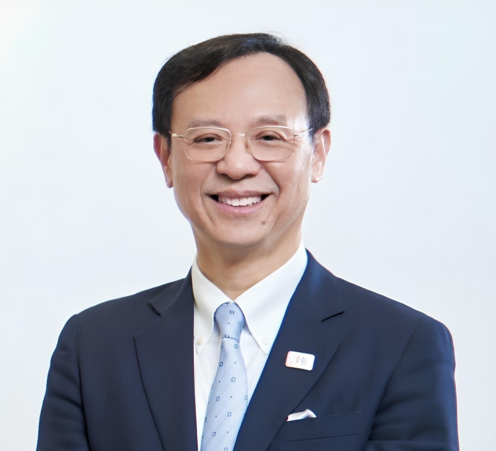 A formal headshot of a smiling man with short dark hair and glasses, wearing a dark navy suit, a white dress shirt, and a light blue patterned tie. He has a small pin on his left lapel and a white pocket square. The background is a solid, bright white.