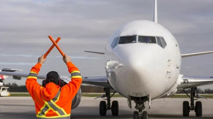 An airport ground crew worker in a high-visibility orange jacket uses crossed wands to guide a large white commercial airplane on the tarmac.